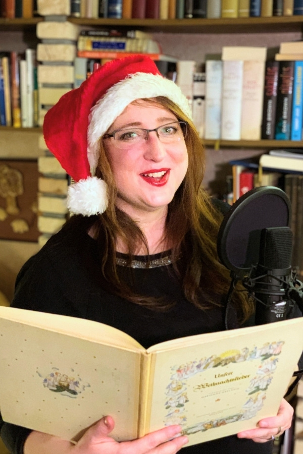 Catrin mit Weihnachtsmannmütze liest vor einer Bücherwand eine Weihnachtsgeschichte ins Mikrofon.